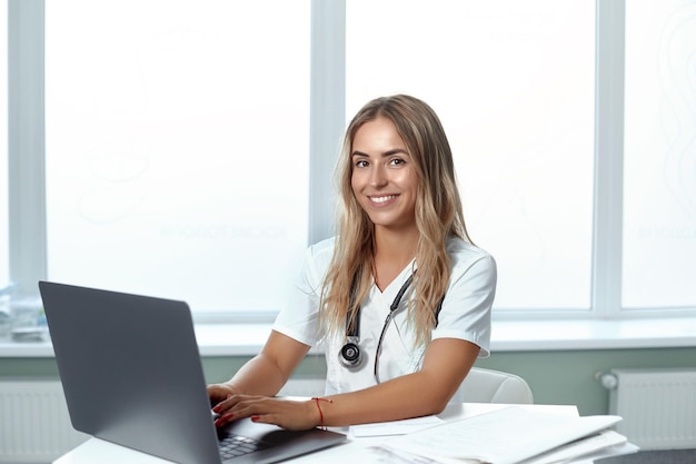 Retrato de una doctora sonriente con una computadora portátil sentada en el escritorio en el consultorio médico Concepto de atención médica