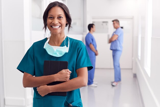 Retrato de doctora sonriente vistiendo matorrales en el pasillo del hospital sosteniendo tableta digital