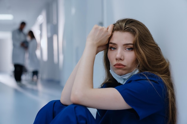 Retrato de una doctora con traje azul sentada en el suelo en el pasillo del hospital y mirando a la cámara