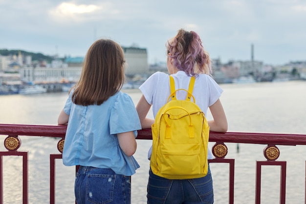 Retrato de dos adolescentes de pie con la espalda en el puente sobre el río, amigas disfrutando de la puesta de sol sobre la superficie del agua, hablando, relajándose. Amistad, estilo de vida, juventud, adolescentes