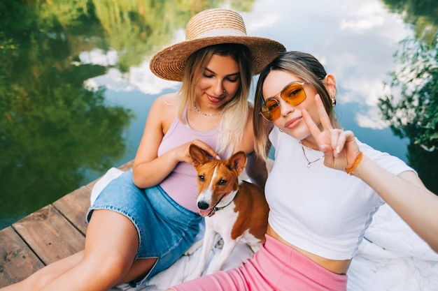 Foto retrato de dos amigas alegres descansando al aire libre en el muelle del lago con perro.