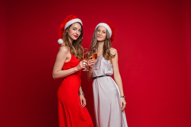 Retrato de dos amigas atractivas con el pelo largo y ondulado en vestidos de seda rojos y blancos posando en sombreros de Santa con copas de vino rosado