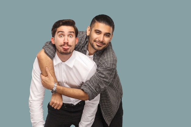 Retrato de dos amigos barbudos felices guapos de pie, disfrutando, abrazándose y mirando a cámara con sonrisa y cara divertida y lengua. tiro del estudio de interior, aislado en fondo azul claro.