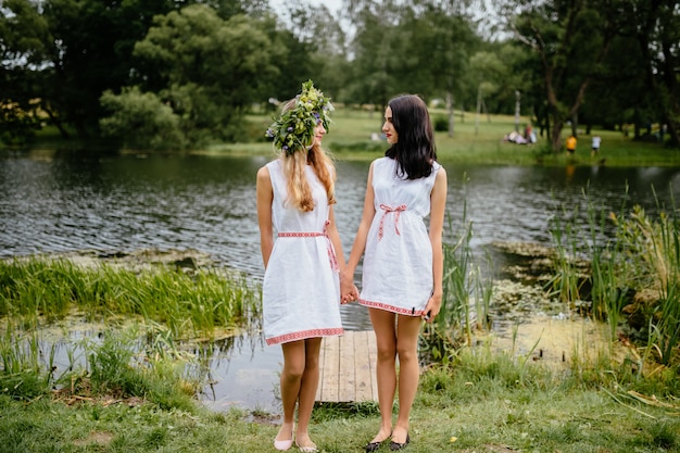 Retrato de dos chicas jóvenes en traje tradicional eslavo con corona de flores de verano. Amigos de estilo étnico folklórico en trajes tradicionales con adornos en la fiesta floral de verano en la naturaleza