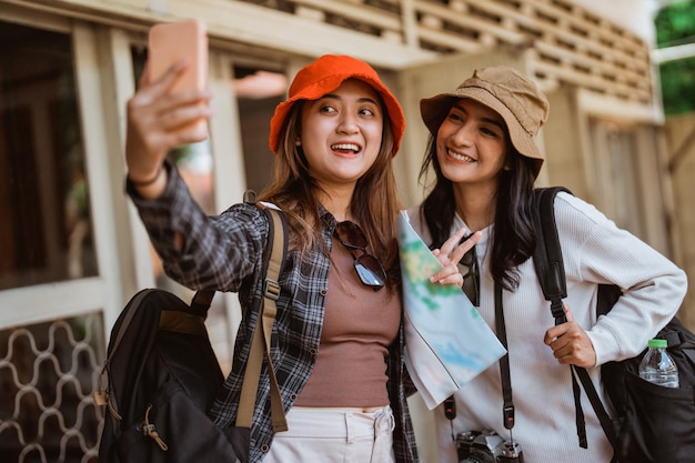 Retrato de dos chicas mochileras haciendo selfies usando un teléfono móvil de pie frente a una casa