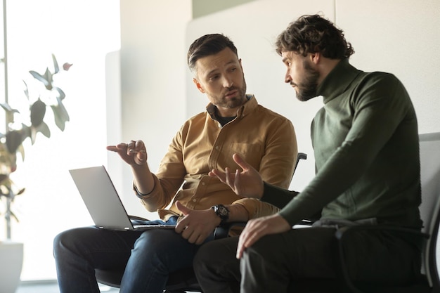 Retrato de dos colegas masculinos discutiendo proyectos de trabajo e ideas de inicio usando una computadora portátil en un moderno