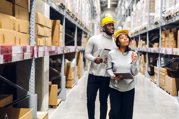 Retrato de dos equipos de ingenieros afroamericanos que envían el detalle de la orden en la tableta, comprueban los bienes y suministros en los estantes con el inventario de mercancías en la industria logística del almacén de la fábrica y la exportación comercial