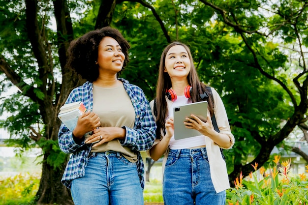 Retrato de dos estudiantes asiáticas y afroamericanas caminando por el parque natural con flores y plantas