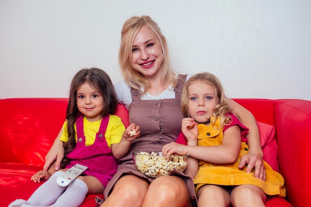 Retrato de dos felices hijas y madres viendo dibujos animados con palomitas de maíz en un sofá rojo