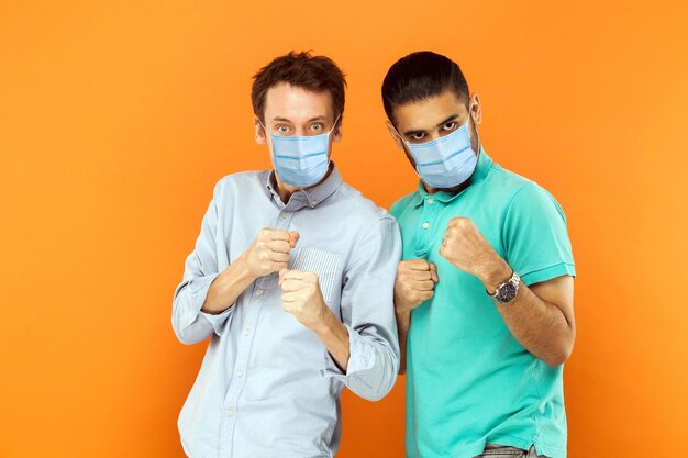 Retrato de dos hombres trabajadores jóvenes serios con máscara médica quirúrgica de pie con puños de boxeo y listos para atacar o defenderse contra virus o problemas. Foto de estudio de interior aislado sobre fondo naranja