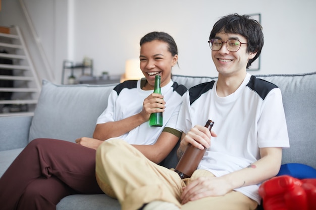 Retrato de dos jóvenes alegres viendo la televisión y bebiendo cerveza en casa, espacio de copia