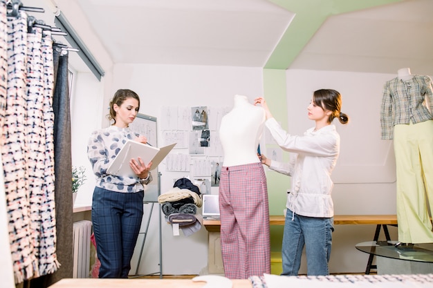 Foto retrato de dos jóvenes diseñadoras de modas o sastres, midiendo maniquí, sosteniendo cinta, trabajando con maniquí en un acogedor estudio de diseño creativo o sastrería, concepto de costura y costura