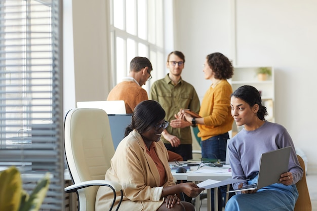 Retrato de dos jóvenes empresarias étnicas discutiendo el proyecto mientras usa la computadora portátil en el interior de la oficina blanca, espacio de copia