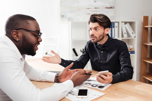 Retrato de dos jóvenes empresarios multinacionales sentados en la mesa y discutiendo emocionalmente un nuevo proyecto mientras trabajan juntos en una oficina aislada