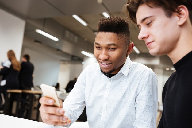 Retrato de dos jóvenes estudiantes alegres sentados en la biblioteca y usando teléfono celular