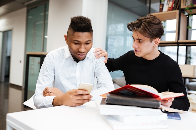 Retrato de dos jóvenes estudiantes sentados en la biblioteca y elegir entre el libro y el teléfono móvil.