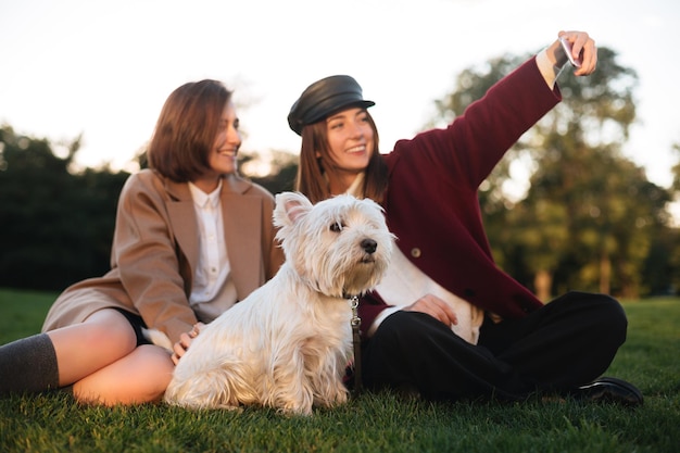 Retrato de dos jóvenes hermosas damas sentadas en el césped y tomándose selfie en el celular mientras pasan tiempo con su lindo perro en el parque