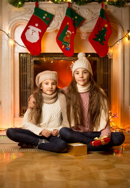 Retrato de dos lindas chicas sentadas junto a la chimenea decorada para Navidad