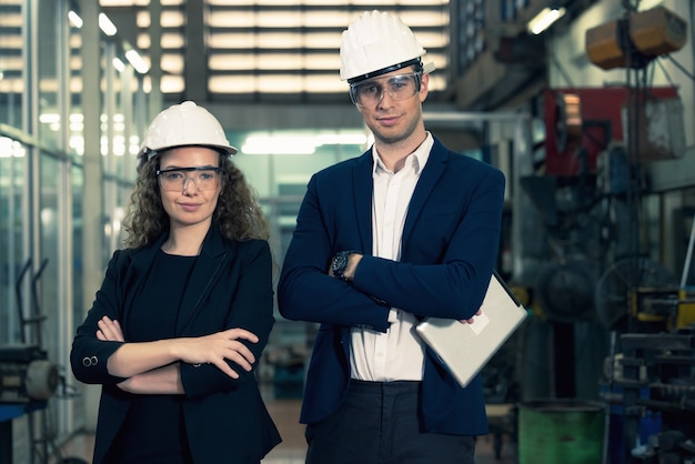 Retrato de dos maquinistas está de pie con confianza con casco de seguridad frente a la pared de vidrio de la fábrica de la industria.