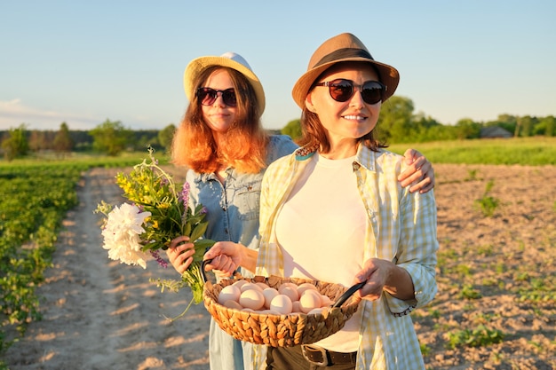 Retrato de dos mujeres abrazadas sonrientes felices madre e hija adolescente con canasta de huevos de granja en sombreros, con ramo de flores en el jardín, naturaleza de verano, fondo puesta de sol