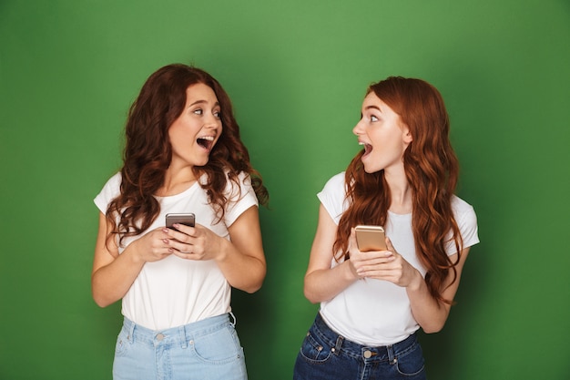 Retrato de dos mujeres adolescentes con cabello jengibre mirando el uno al otro y usando teléfonos celulares, aislado sobre fondo verde