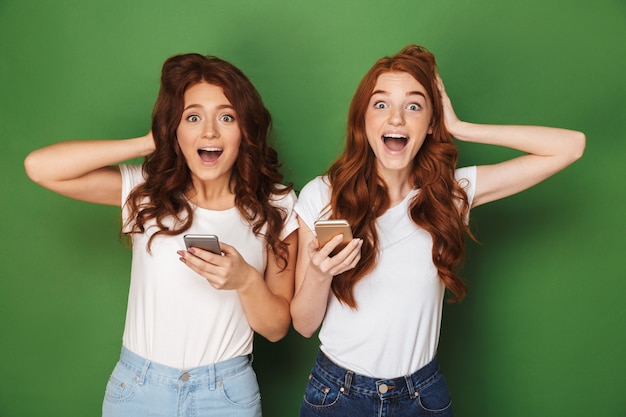 Retrato de dos mujeres alegres con cabello jengibre mirando a la cámara y sosteniendo teléfonos móviles, aislado sobre fondo verde