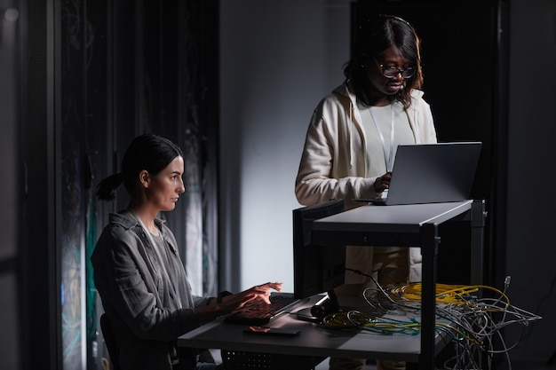 Retrato de dos mujeres ingeniero de redes con ordenador portátil mientras trabaja en la sala de servidores oscura, espacio de copia