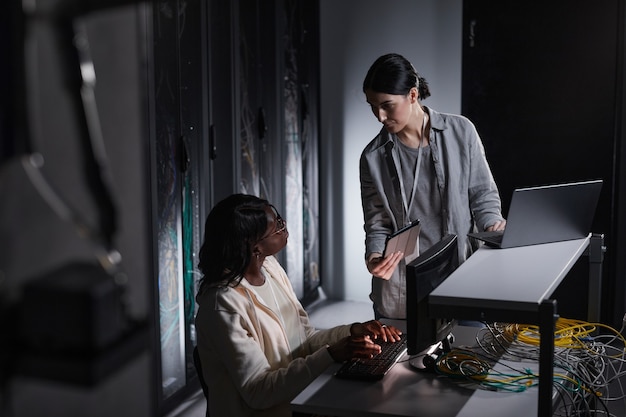 Retrato de dos mujeres ingeniero de redes usando laptop mientras trabajan juntos en la sala de servidores oscura, espacio de copia