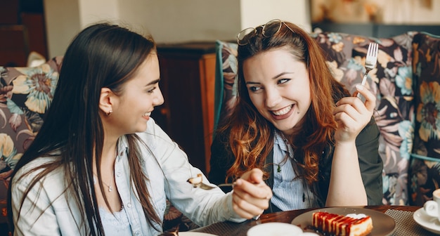 Retrato de dos mujeres jóvenes encantadoras, contando historias y comiendo pastel de queso y riendo en una cafetería.