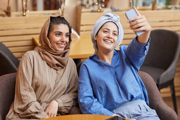 Retrato de dos mujeres jóvenes del Medio Oriente tomando selfie a través de un teléfono inteligente mientras se relaja en la cafetería, espacio de copia