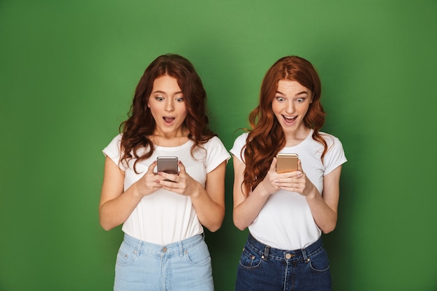 Retrato de dos mujeres pelirrojas emocionadas de 20 años posando en la cámara y usando teléfonos inteligentes, aislado sobre fondo verde