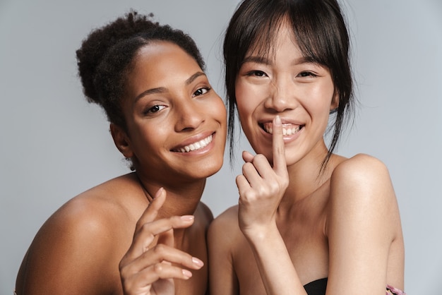 Foto retrato de dos mujeres semidesnudas multinacionales haciendo gesto de silencio y sonriendo