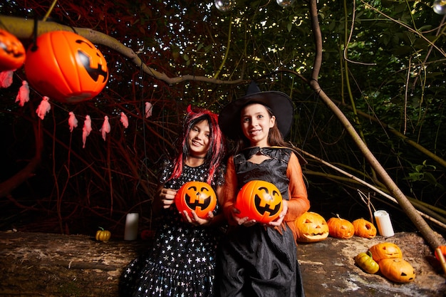 Retrato de dos niñas en disfraces sosteniendo calabazas de juguete y sonriendo a la cámara están en la fiesta en la oscuridad