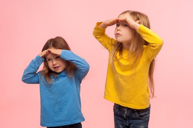 Retrato de dos niñas graciosas y divertidas cogidas de la mano sobre los ojos y mirando a lo lejos con una curiosa expresión atenta buscando una distancia de visualización en un estudio interior aislado en un fondo rosa