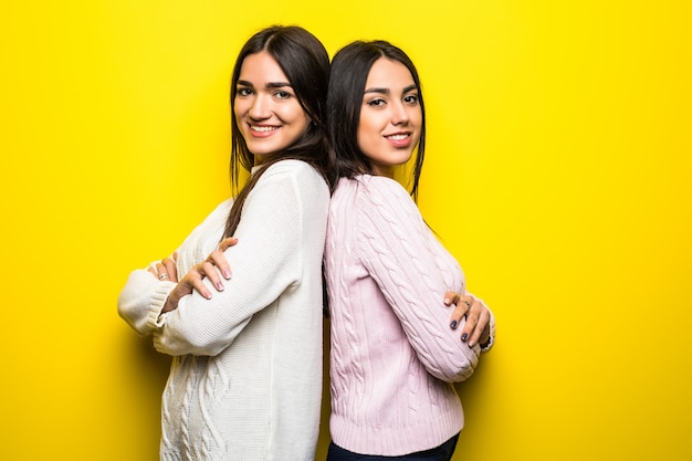 Retrato de dos niñas sonrientes vestidas con suéteres de pie espalda con espalda aislado sobre pared amarilla