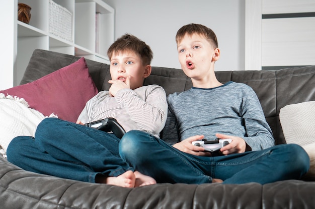 Retrato de dos niños adolescentes conmocionados sentados en un sofá gris en casa con la boca abierta sosteniendo el controlador de juegos