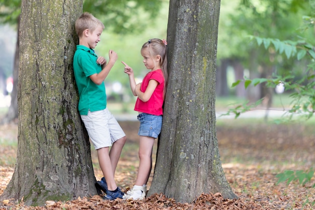 Retrato de dos niños muy lindos niño y niña de pie cerca del tronco de un árbol grande en el parque de verano al aire libre.