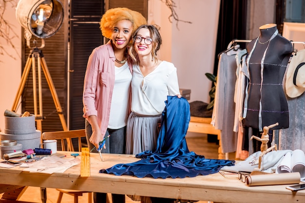 Retrato de dos sastres multiétnicos que trabajan con tela azul en el estudio con diferentes herramientas y ropa de sastrería