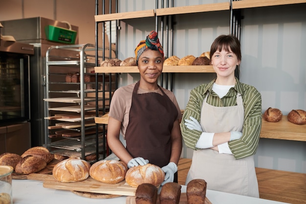 Retrato de dos vendedoras en delantales de pie cerca de la mesa con pan casero en la panadería