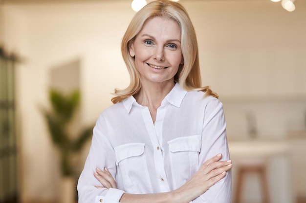 Retrato de elegancia de amable mujer madura con camisa blanca sonriendo a la cámara mientras posa en interiores