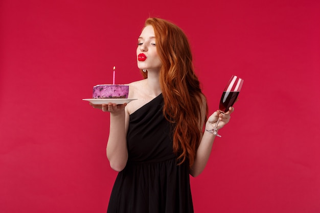 Retrato de una elegante y atractiva mujer pelirroja femenina con lápiz labial rojo, maquillaje de noche y vestido negro, celebrando el cumpleaños en la fiesta con vino tinto y pastel de cumpleaños, soplando velas sensualmente