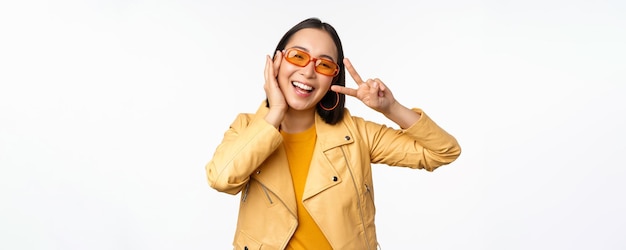 Retrato de una elegante chica asiática moderna con gafas de sol y una chaqueta amarilla que muestra un gesto de paz frente a un rostro sonriente feliz de fondo blanco