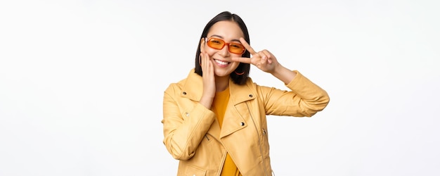 Retrato de una elegante chica asiática moderna con gafas de sol y una chaqueta amarilla que muestra un gesto de paz vs.