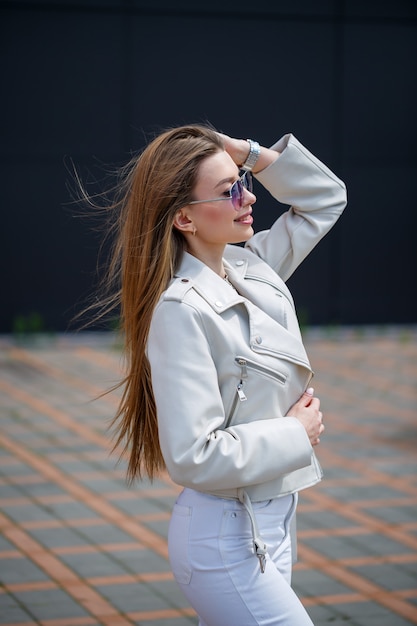 Retrato de una elegante chica de pelo largo con una blusa blanca y pantalones vaqueros ligeros está parado con una sonrisa en el contexto de una pared gris de un edificio en un día soleado de primavera.