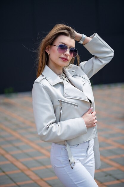 Retrato de una elegante chica de pelo largo con una blusa blanca y pantalones vaqueros ligeros está parado con una sonrisa en el contexto de una pared gris de un edificio en un día soleado de primavera.