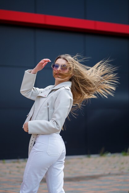 Retrato de una elegante chica de pelo largo con una blusa blanca y pantalones vaqueros ligeros está parado con una sonrisa en el contexto de una pared gris de un edificio en un día soleado de primavera.