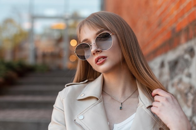 Retrato elegante femenino urbano de una hermosa mujer de moda con gafas de sol hipster redondas vintage en una chaqueta de cuero se sienta cerca de un centro comercial vintage en la calle