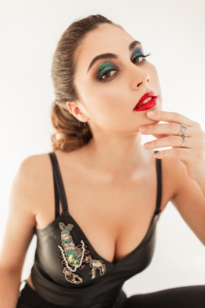 Retrato elegante de una hermosa joven modelo sexy con maquillaje profesional, labios rojos y sombras verdes en un look moderno con una parte superior de cuero y pechos sentados sobre un fondo blanco