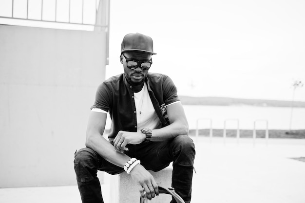 Retrato de un elegante hombre afroamericano sentado con gafas de sol y gorra mirando sus relojes al aire libre Street fashion black man