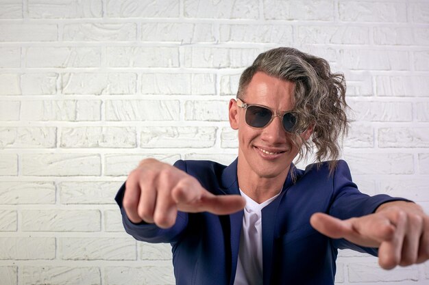 Retrato de un elegante hombre de negocios con el pelo largo y rizado con gafas de sol sonriendo a la cámara y gesticulando con un signo de excelencia en el fondo blanco